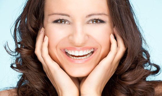 Close-up picture of a smiling woman with long dark hair and perfect teeth, looking directly at the camera with both hands to the side of her face, showing her happiness with the implant-supported bridges procedure she had at Premier Holistic Dental in beautiful Costa Rica.