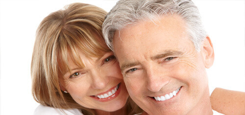 Close-up picture of a smiling couple looking directly into the camera showing their happiness with  having holistic dental work done in Costa Rica.  The woman has medium brown hair and the man has white hair.